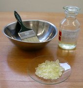 An image showing a paint brush in a metal bowl, a bottle of acid and a plate of beige solid crystals
