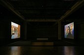 two film projections face each other in a dark space, one of the screens shows a person in a burka, the other screen shows a stone building