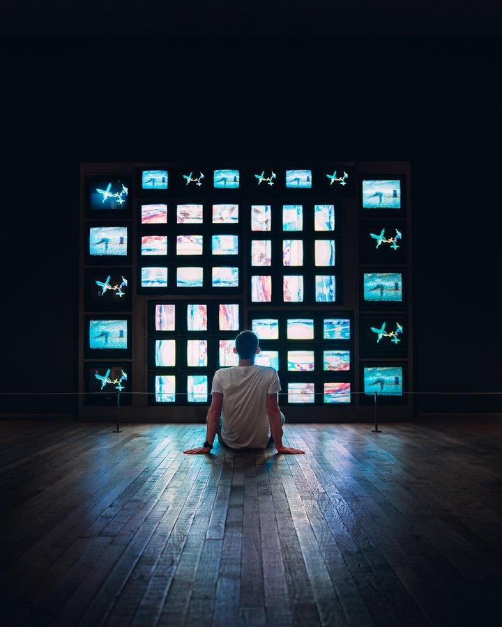 person sat in dark room on floor in front of wall of tv sets