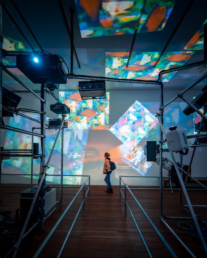 person in gallery surrounded by a metal structure and projections on the walls